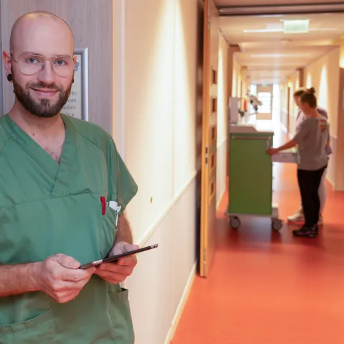 Een zorgmedewerker in groen uniform staat met een tablet in de hand, terwijl collega’s zich in de achtergrond bezighouden met zorgtaken bij ViVa! Zorggroep.